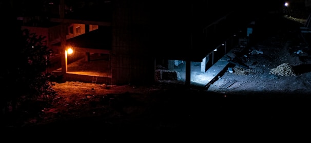 A dimly lit construction site with an orange streetlight illuminating part of the ground and building. The area under construction appears mostly dark, with some scattered rubble and materials. There is a mix of both shadowed and faintly lit areas, adding to the mood of an incomplete, deserted space.