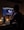 A person is sitting at a desk, looking at a computer monitor displaying video editing software. The room is dimly lit, with warm lighting from the monitor and a smartphone on the desk. A water bottle and small plants are also visible on the desk.