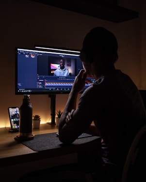A person is sitting at a desk, looking at a computer monitor displaying video editing software. The room is dimly lit, with warm lighting from the monitor and a smartphone on the desk. A water bottle and small plants are also visible on the desk.
