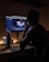 A person is sitting at a desk, looking at a computer monitor displaying video editing software. The room is dimly lit, with warm lighting from the monitor and a smartphone on the desk. A water bottle and small plants are also visible on the desk.