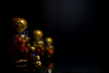 A row of Matryoshka dolls is featured, with the smallest doll prominently in focus at the front. The dolls are intricately decorated with bright floral patterns, featuring colors like blue, red, and gold. The lighting is low, creating a dramatic, shadowy effect with a dark background, emphasizing the detail and color of the dolls.