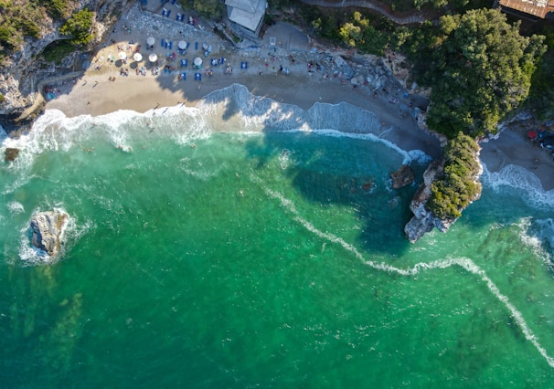 An overhead view of a coastal beach with turquoise waves gently rolling onto white sand, dotted with colorful umbrellas.