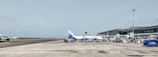 A busy airport apron featuring a passenger aircraft parked near the terminal with boarding bridges and ground service equipment surrounding it. The plane has the Indigo logo on its tail. The terminal building is visible on the right side with multiple glass windows. The scene includes clear skies and some airport vehicles nearby.