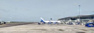 A busy airport apron featuring a passenger aircraft parked near the terminal with boarding bridges and ground service equipment surrounding it. The plane has the Indigo logo on its tail. The terminal building is visible on the right side with multiple glass windows. The scene includes clear skies and some airport vehicles nearby.