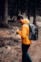 A person wearing an orange jacket and carrying a backpack stands in a forested area, focusing on a camera in their hands. Tall trees and a dirt path are visible in the background, and the scene suggests a calm and natural environment.