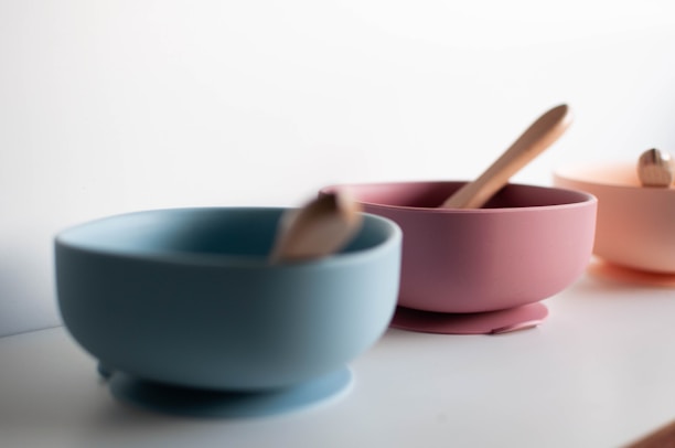 Three pastel-colored bowls, each with a wooden spoon inside, are arranged in a row on a smooth white surface. The bowls are in shades of blue, pink, and peach, with a minimalist and modern design.