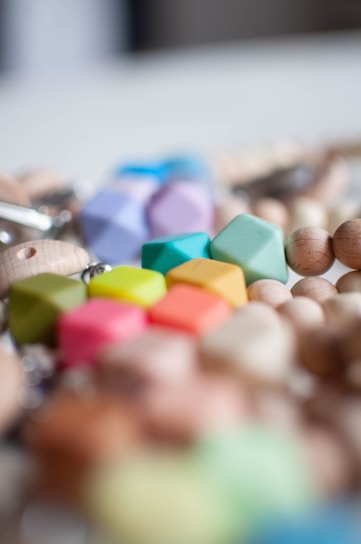 A close-up view of colorful geometric beads made from various materials, including wood and plastic. The beads are arranged in a scattered manner, displaying hues of pink, green, blue, yellow, orange, and purple, with some unfinished wooden beads. The image is slightly blurred in the background, bringing focus to the vivid colors and shapes in the foreground.