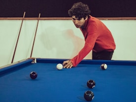 A person wearing a red sweater leans over a pool table, preparing to take a shot with the white cue ball. Several black balls are scattered across the blue felt surface of the table. Cue sticks are resting against the wall in the background.