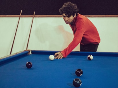 A person wearing a red sweater leans over a pool table, preparing to take a shot with the white cue ball. Several black balls are scattered across the blue felt surface of the table. Cue sticks are resting against the wall in the background.