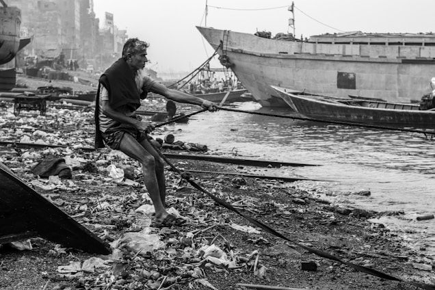 A person is sitting by a polluted riverbank, pulling on a rope attached to a boat. The area is cluttered with debris and litter. Large wooden boats are docked nearby, and buildings are visible in the background through a hazy atmosphere.