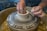 Hands expertly shape a piece of clay on a pottery wheel, with water and clay drips visible, indicating active crafting. The scene suggests a focus on creating ceramic art.