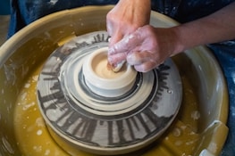 Hands are shaping a clay pot on a pottery wheel, capturing the intricate process of pottery making. The wheel spins as the artisan's fingers carefully mold the clay into a symmetrical form.