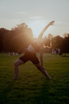 A person is performing a yoga pose on a grassy field with the sun setting in the background. The warm sunlight creates a silhouette effect against the individual. There are trees in the distance and people visible in the background, adding to the peaceful atmosphere.
