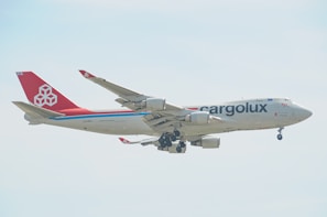 A large cargo airplane with the 'Cargolux' branding is captured mid-flight against a light, slightly cloudy sky. The aircraft has a predominantly white fuselage with a distinct red tail and engines. The branding and company logo are visible, along with the registration code on the side.