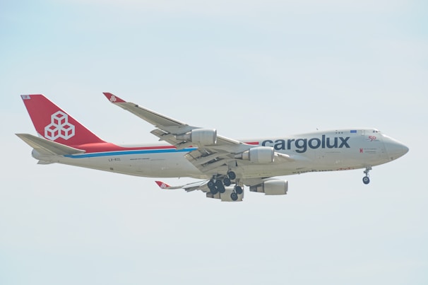 A large cargo airplane with the 'Cargolux' branding is captured mid-flight against a light, slightly cloudy sky. The aircraft has a predominantly white fuselage with a distinct red tail and engines. The branding and company logo are visible, along with the registration code on the side.