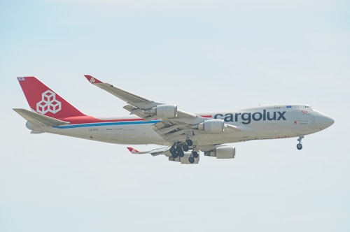 A large cargo airplane with the 'Cargolux' branding is captured mid-flight against a light, slightly cloudy sky. The aircraft has a predominantly white fuselage with a distinct red tail and engines. The branding and company logo are visible, along with the registration code on the side.