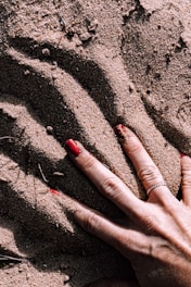 A hand with red nail polish is pressing down on textured sand, creating visible grooves and patterns. The hand is adorned with a silver ring.