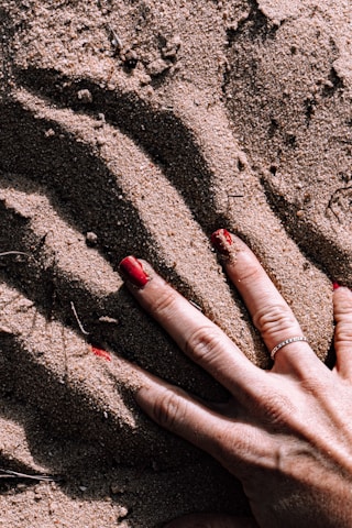 A hand with red nail polish is pressing down on textured sand, creating visible grooves and patterns. The hand is adorned with a silver ring.