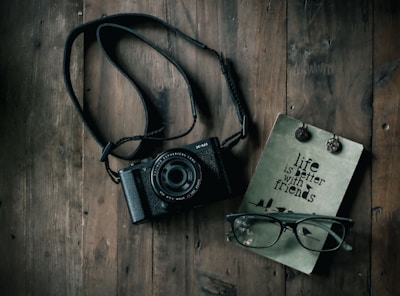 A black camera with a strap lies on a rustic wooden surface alongside a pair of eyeglasses and a journal with the text 'life is better with friends.' Two decorative buttons or pins are attached to the journal.
