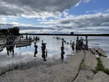 An array of rock sculptures is artfully balanced along a water's edge, with their reflections creating a mirror-like effect on the water's surface. The scene is set under a dynamic sky filled with fluffy clouds, where patches of blue sky peek through. The horizon shows distant trees and a faint silhouette of a building, indicating a serene outdoor setting.