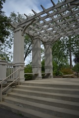 A wooden pergola structure with large beams and columns is surrounded by greenery. The pergola has open slats at the top, allowing sunlight to filter through. Concrete steps lead up to the pergola, and metal railings are fixed to the sides. Trees and bushes fill the background.