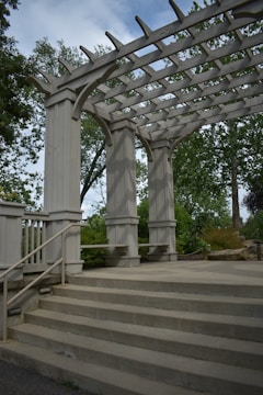A wooden pergola structure with large beams and columns is surrounded by greenery. The pergola has open slats at the top, allowing sunlight to filter through. Concrete steps lead up to the pergola, and metal railings are fixed to the sides. Trees and bushes fill the background.