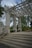 A wooden pergola structure with large beams and columns is surrounded by greenery. The pergola has open slats at the top, allowing sunlight to filter through. Concrete steps lead up to the pergola, and metal railings are fixed to the sides. Trees and bushes fill the background.