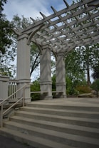 A wooden pergola structure with large beams and columns is surrounded by greenery. The pergola has open slats at the top, allowing sunlight to filter through. Concrete steps lead up to the pergola, and metal railings are fixed to the sides. Trees and bushes fill the background.