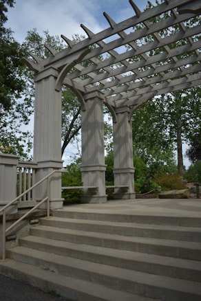 A wooden pergola structure with large beams and columns is surrounded by greenery. The pergola has open slats at the top, allowing sunlight to filter through. Concrete steps lead up to the pergola, and metal railings are fixed to the sides. Trees and bushes fill the background.