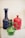 Ornate glass bottles are displayed, featuring intricate designs and vibrant colors. A prominent green glass pitcher with a twisted handle stands in the foreground, accompanied by a tall blue bottle with a decorative pattern and a red bottle with a molded design in the background.