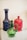 Ornate glass bottles are displayed, featuring intricate designs and vibrant colors. A prominent green glass pitcher with a twisted handle stands in the foreground, accompanied by a tall blue bottle with a decorative pattern and a red bottle with a molded design in the background.