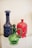 Ornate glass bottles are displayed, featuring intricate designs and vibrant colors. A prominent green glass pitcher with a twisted handle stands in the foreground, accompanied by a tall blue bottle with a decorative pattern and a red bottle with a molded design in the background.