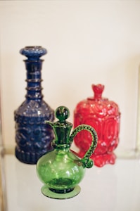 Ornate glass bottles are displayed, featuring intricate designs and vibrant colors. A prominent green glass pitcher with a twisted handle stands in the foreground, accompanied by a tall blue bottle with a decorative pattern and a red bottle with a molded design in the background.
