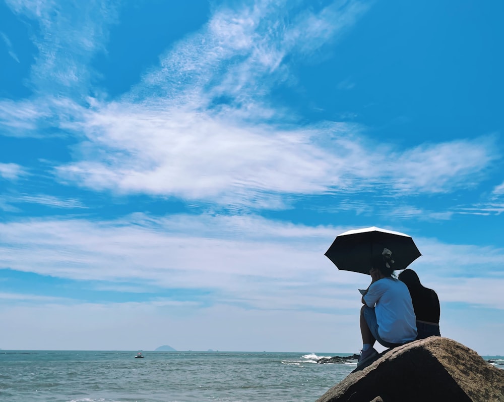person in black hat sitting on rock near sea under blue sky during daytime