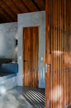A minimalist interior featuring a wooden door and adjacent wooden screen panel. The walls and floor are polished concrete, and a built-in seating area with a dark cushion is partially visible. Sunlight casts shadows through the wooden slats.