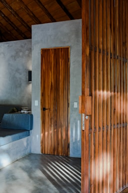A minimalist interior featuring a wooden door and adjacent wooden screen panel. The walls and floor are polished concrete, and a built-in seating area with a dark cushion is partially visible. Sunlight casts shadows through the wooden slats.