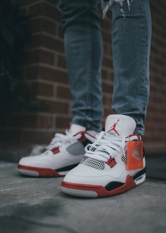 A pair of white and red sneakers with black accents is worn by a person standing on a pavement. The jeans are slightly distressed, and a brick wall is in the background. A tag with the Nike Air logo dangles from the left shoe.