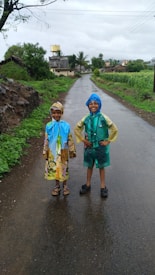 Two children wearing colorful raincoats stand smiling on a wet village road. The surrounding area is lush with greenery, and there are small houses and a water tank in the distance under a cloudy sky.
