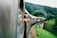 A train traveling through a lush green landscape with passengers leaning out of the windows, their arms extended into the open air. The train curves along a track over a bridge, with rolling hills and dense forests in the background.