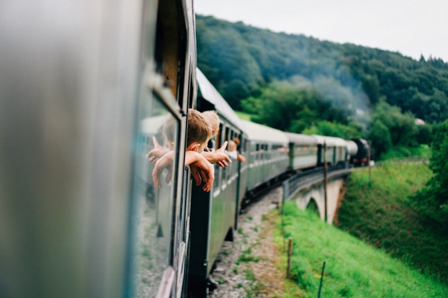 A train traveling through a lush green landscape with passengers leaning out of the windows, their arms extended into the open air. The train curves along a track over a bridge, with rolling hills and dense forests in the background.