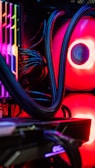 A close-up view of the interior of a computer tower showcasing various components. Brightly colored LED lights illuminate the cables, cooling pipes, and heatsinks, creating a vibrant display of red and other colors against a dark background. The composition highlights the intricate wiring and hardware setup.