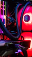A close-up view of the interior of a computer tower showcasing various components. Brightly colored LED lights illuminate the cables, cooling pipes, and heatsinks, creating a vibrant display of red and other colors against a dark background. The composition highlights the intricate wiring and hardware setup.