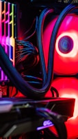 A close-up view of the interior of a computer tower showcasing various components. Brightly colored LED lights illuminate the cables, cooling pipes, and heatsinks, creating a vibrant display of red and other colors against a dark background. The composition highlights the intricate wiring and hardware setup.