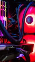 A close-up view of the interior of a computer tower showcasing various components. Brightly colored LED lights illuminate the cables, cooling pipes, and heatsinks, creating a vibrant display of red and other colors against a dark background. The composition highlights the intricate wiring and hardware setup.