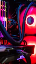 A close-up view of the interior of a computer tower showcasing various components. Brightly colored LED lights illuminate the cables, cooling pipes, and heatsinks, creating a vibrant display of red and other colors against a dark background. The composition highlights the intricate wiring and hardware setup.