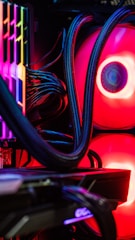 A close-up view of the interior of a computer tower showcasing various components. Brightly colored LED lights illuminate the cables, cooling pipes, and heatsinks, creating a vibrant display of red and other colors against a dark background. The composition highlights the intricate wiring and hardware setup.