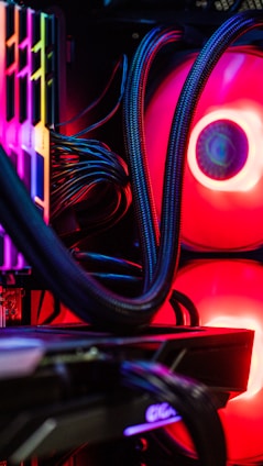 A close-up view of the interior of a computer tower showcasing various components. Brightly colored LED lights illuminate the cables, cooling pipes, and heatsinks, creating a vibrant display of red and other colors against a dark background. The composition highlights the intricate wiring and hardware setup.