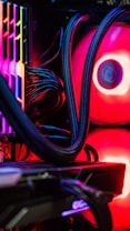 A close-up view of the interior of a computer tower showcasing various components. Brightly colored LED lights illuminate the cables, cooling pipes, and heatsinks, creating a vibrant display of red and other colors against a dark background. The composition highlights the intricate wiring and hardware setup.