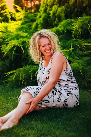 A woman with curly blonde hair is sitting on grass, smiling and looking at her knee while wearing a sleeveless dress adorned with abstract black text on a light background. Dense green foliage forms a lush backdrop.