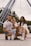 A family of three is posing together outdoors in front of a large Ferris wheel. The father, wearing a white shirt and denim shorts, crouches beside the mother, who is dressed in white and wearing glasses. Their young child stands between them pointing forward, smiling slightly. The setting appears to be a park or amusement area, with trees in the background.